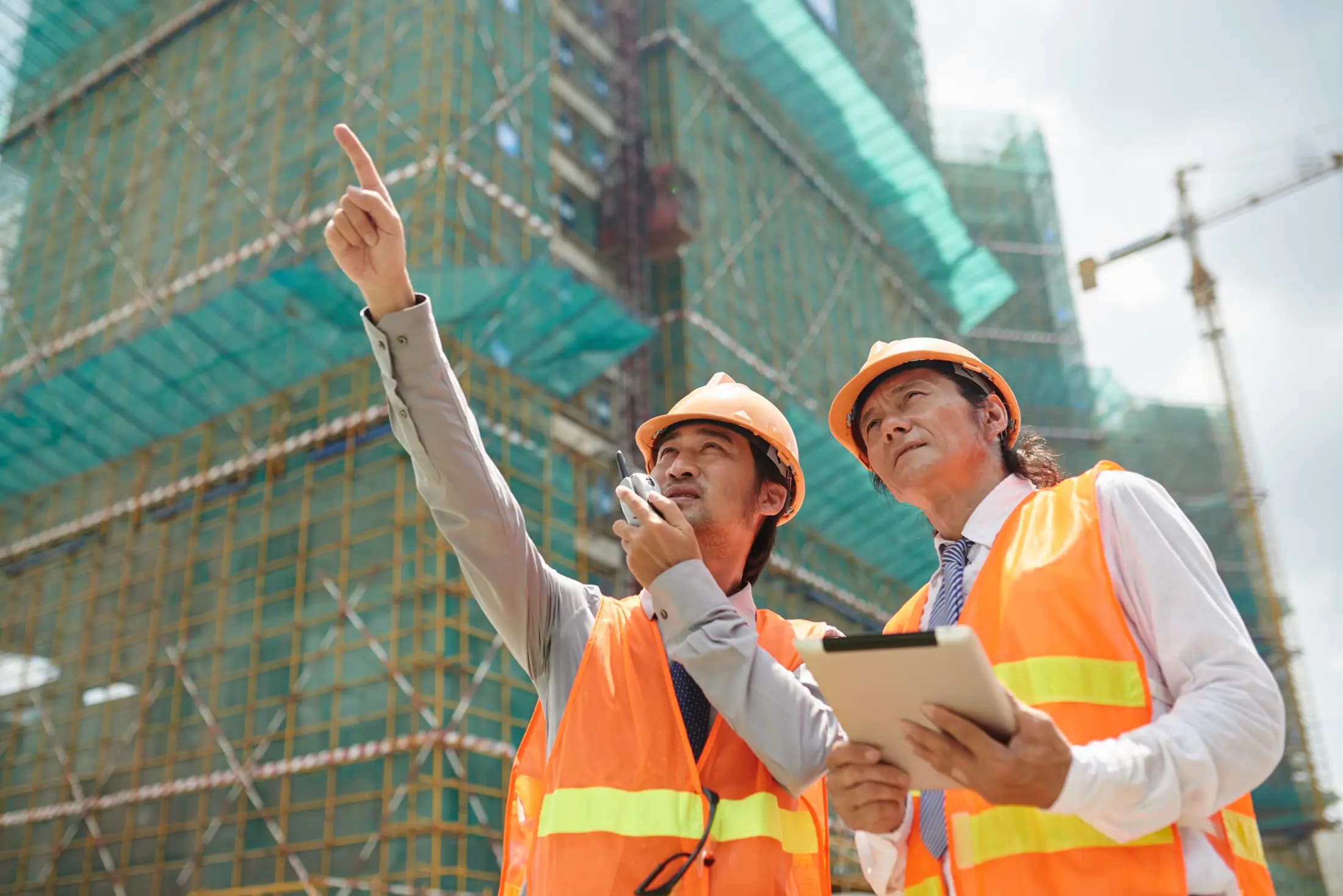 Two site engineers in hard hats and safety vests reviewing a high-rise construction site
